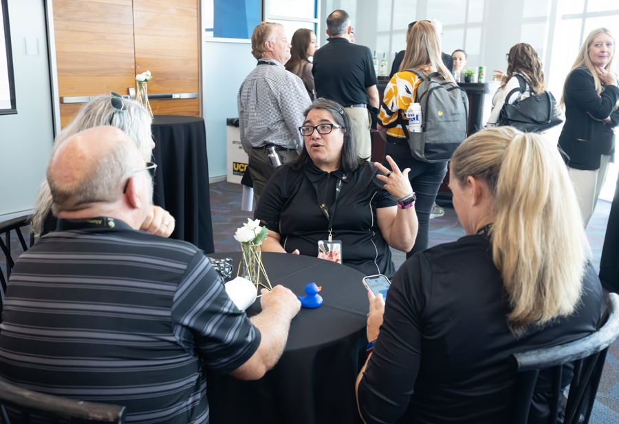 Professionals at a round-table networking session, a woman in black speaks and gestures as others listen nearby at a conference venue.]','Professionals networking at a conference lounge, with attendees standing in the background while a woman wearing glasses talks at a small round table.