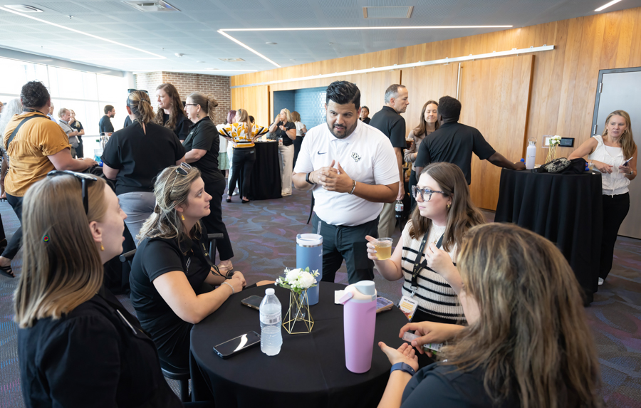 Man in a white polo speaks with four women seated around a round table at a networking event.
