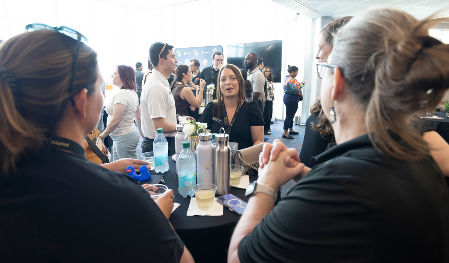 Professionals mingle around a high round table at a conference networking event with drinks and a small centerpiece in the middle by a blue backdrop.