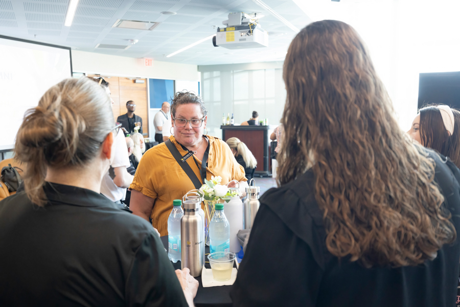 Woman in a mustard-yellow shirt talks with others around a refreshment table at a conference or workshop venue.