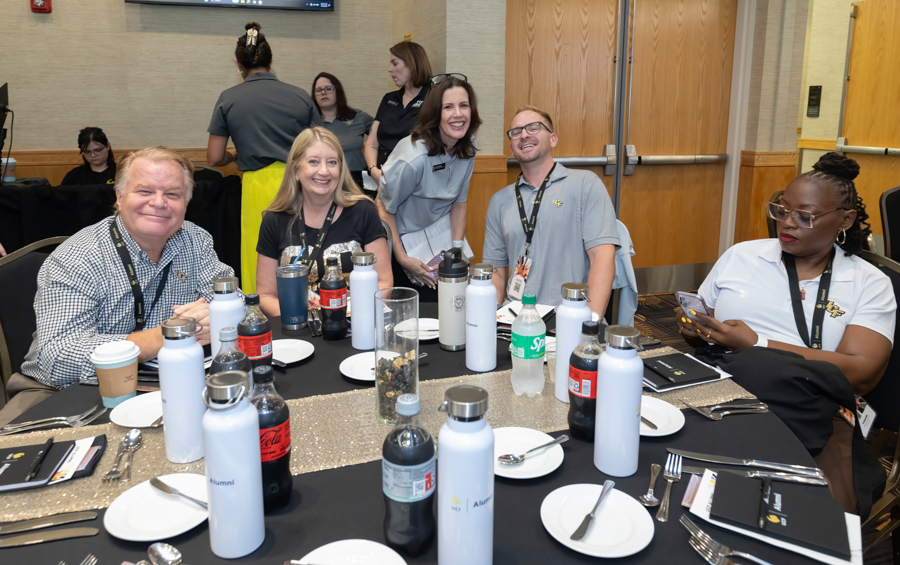 Group of adults seated around a round banquet table at a conference, smiling for the camera with drinks and plates nearby.