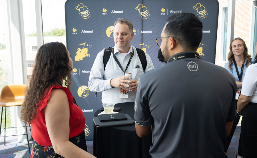 Group of people networking at a university alumni event near a branded backdrop with UCF Alumni logos and mascots.