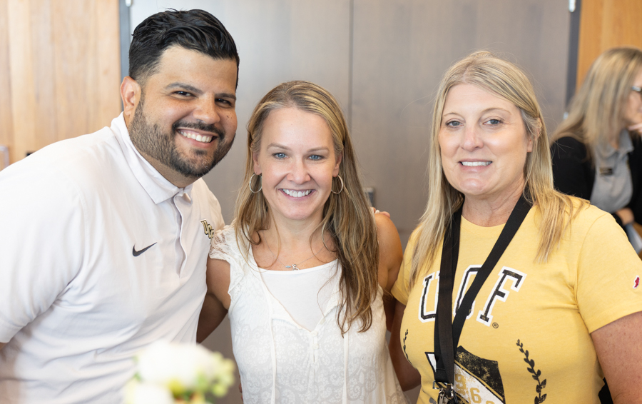 Three adults posing and smiling at an indoor event; man in white polo, woman in white top, woman in yellow UF shirt with a lanyard.