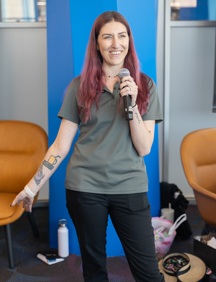 Woman with pink-tinted hair holding a microphone, speaking indoors in front of a blue backdrop.