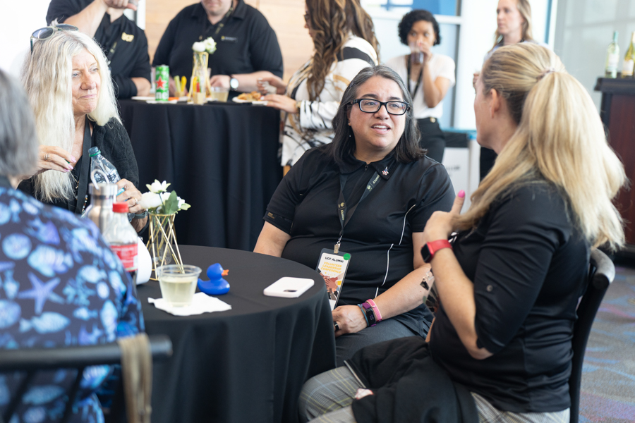 Woman with glasses at a round table chats with a colleague during a networking event; others mingle in the background.