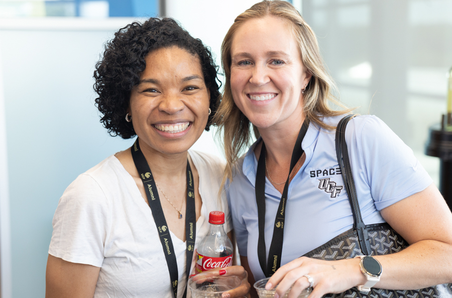 Two smiling women with lanyards pose together at a social event, one holding a Coca-Cola bottle and a cup of drink.