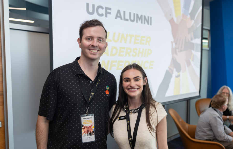 Two smiling attendees with lanyards posing for a photo at a UCF Alumni Volunteer Leadership Conference, with a presentation screen behind them.