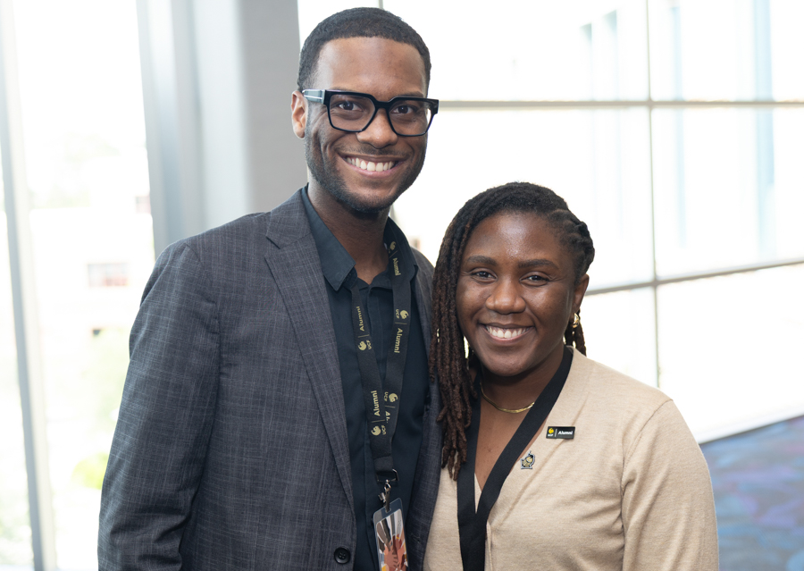 Two smiling professionals pose together in a bright office lobby, wearing lanyards and badges.