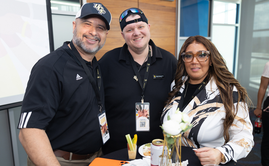 Three smiling adults pose together at a casual event, wearing lanyards and name badges near a table with drinks and flowers.