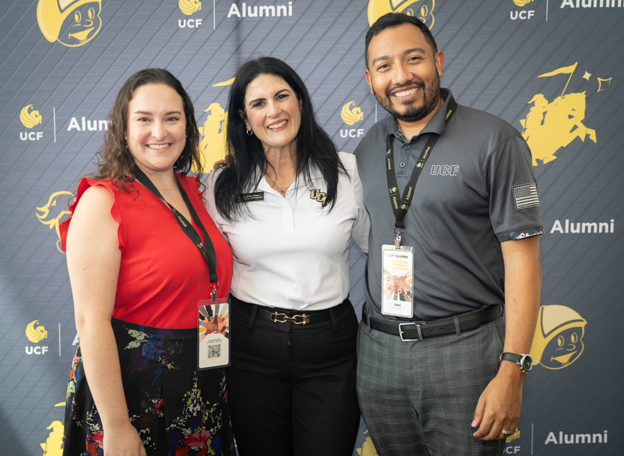 Three smiling adults at a UCF Alumni event pose in front of a branded backdrop; left in a red top, center in a white polo, right in a gray polo with badge.