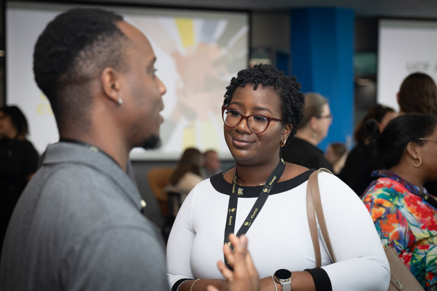 Two people converse at a networking event; woman with red glasses and lanyard speaks to a man in a gray blazer, others mingling in background.