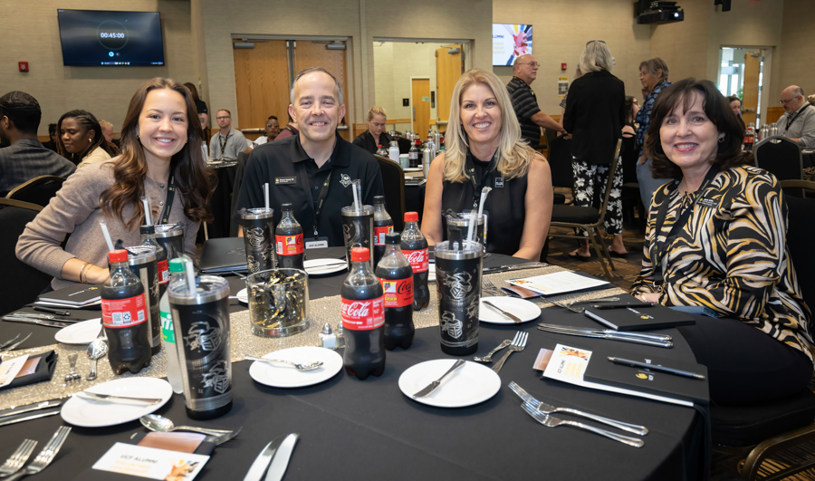 Group of four adults smiling at a round banquet table during an indoor event, with assorted drinks and place settings.