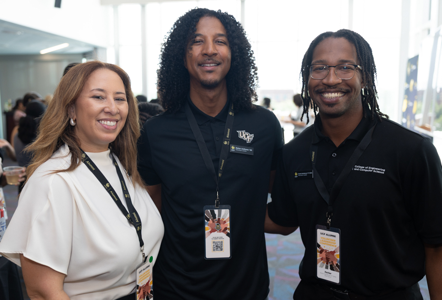 Three smiling attendees posing together at a bright event space, wearing lanyards and badges.