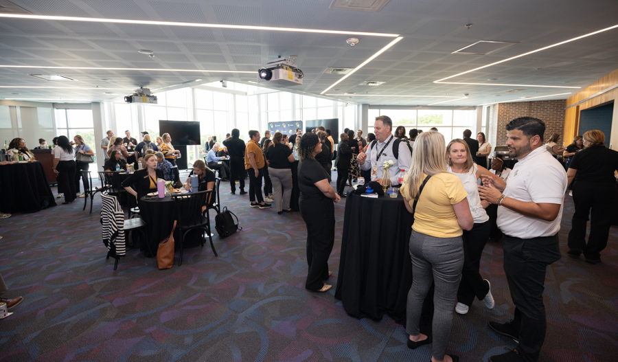 Networking event in a bright conference room with groups chatting around high-top tables and a projector screen in the background.