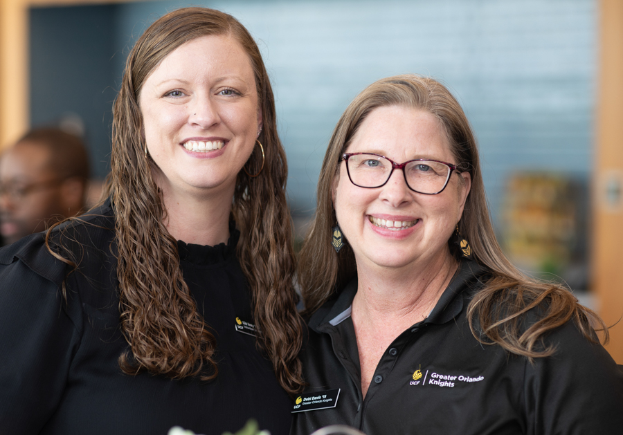 Two women in black polo shirts smile for a photo at an event, standing close together with a blurred background behind them.