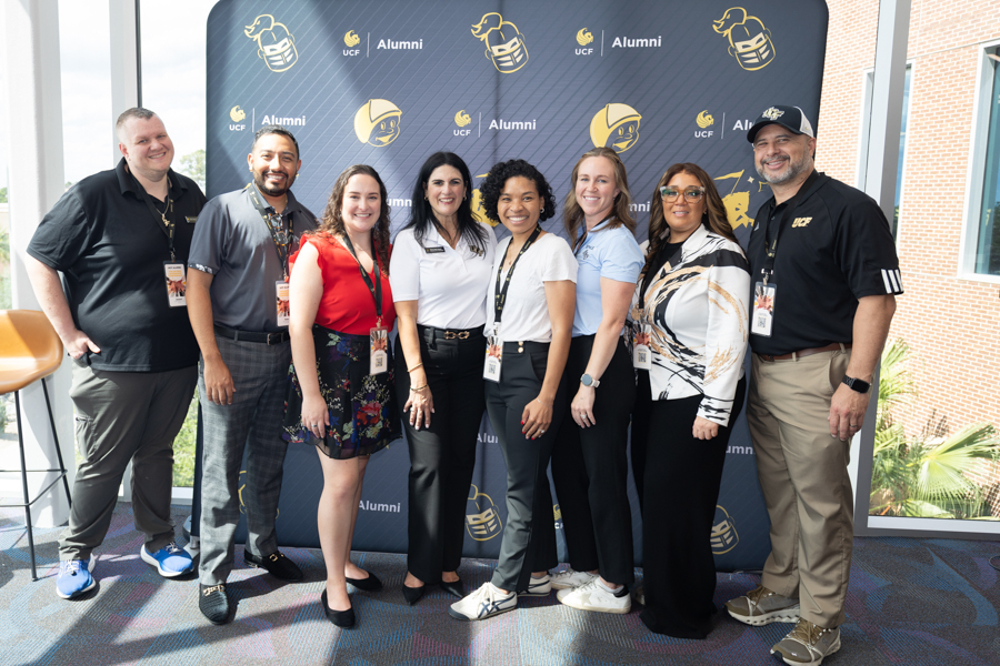 Nine adults pose for a group photo at a UCF Alumni event in front of a branded backdrop with UCF Alumni logos, all wearing event lanyards and badges.