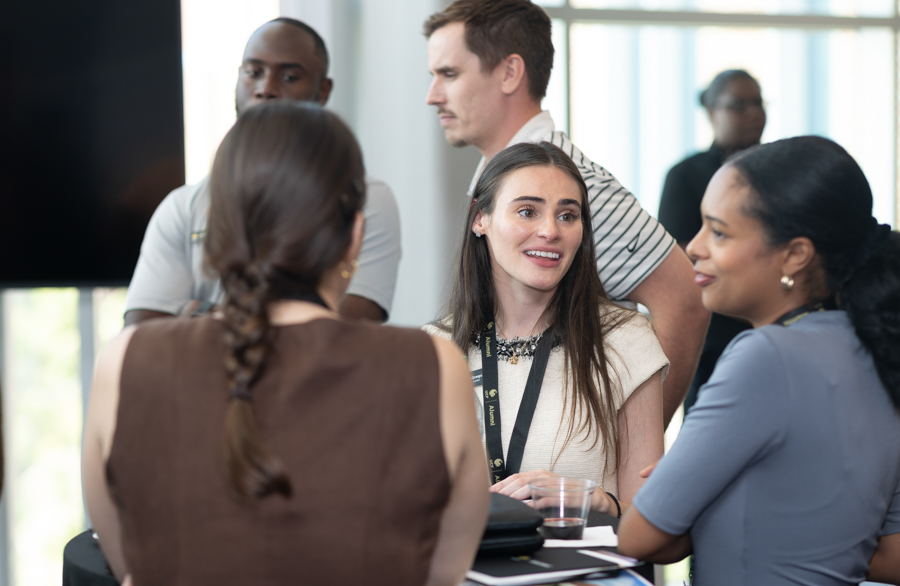 Group of professionals chatting at a networking event around a high table; woman with lanyard smiles at the center.