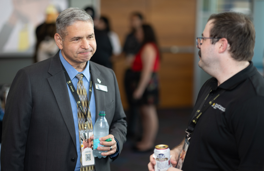 Two men conversing at a conference: one in a suit holding a water bottle, the other in a black polo with a can, blurred attendees in the background.