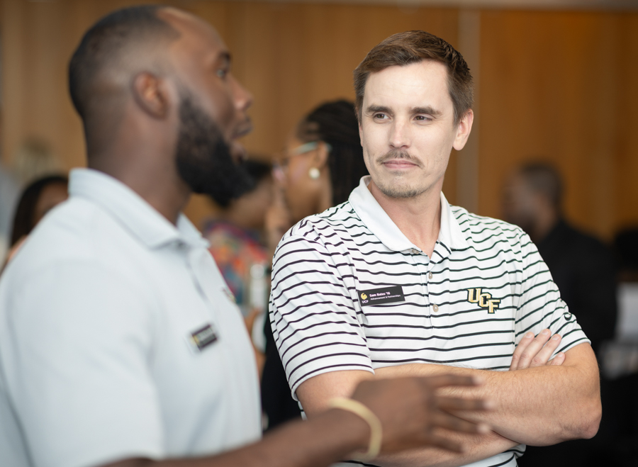 Two men converse at a networking event; the man on the right wears a striped polo with a UCF logo and crossed arms.