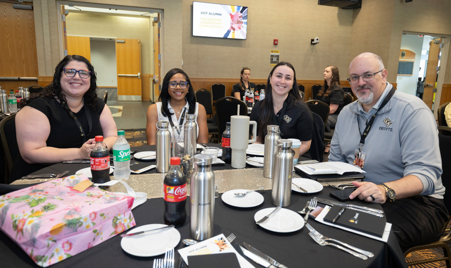 Four attendees at a conference table smiling for the camera; drinks, plates, and a pink gift bag on the table, with a conference screen in the background.