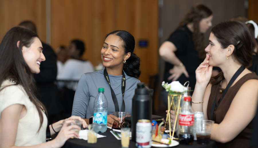 Three women sit around a table at a social event, smiling and chatting with drinks and snacks on the table.”] ,