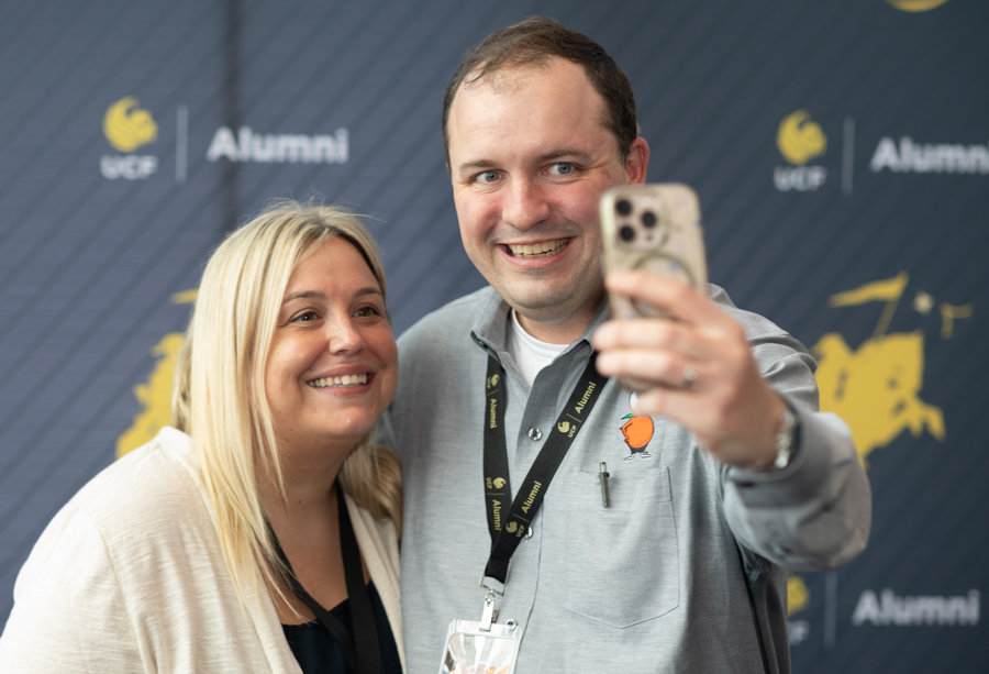 Two smiling adults take a selfie at an alumni event, with a UCF Alumni backdrop in the background.