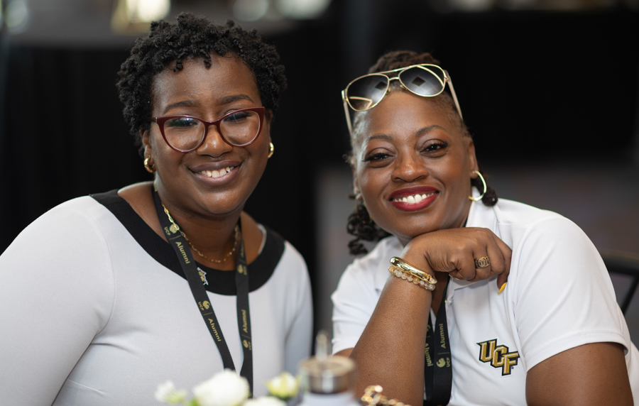 Two women smiling at a social event, wearing lanyards and white tops with a UCF logo on one shirt.