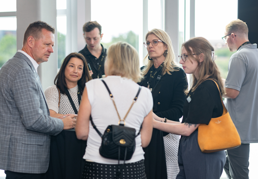 Group of professionals conversing at a networking event beside large windows in a bright space.