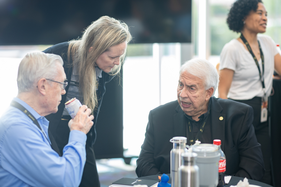 Two older men and a woman converse at a conference table; one man drinks from a can while listening to the others.