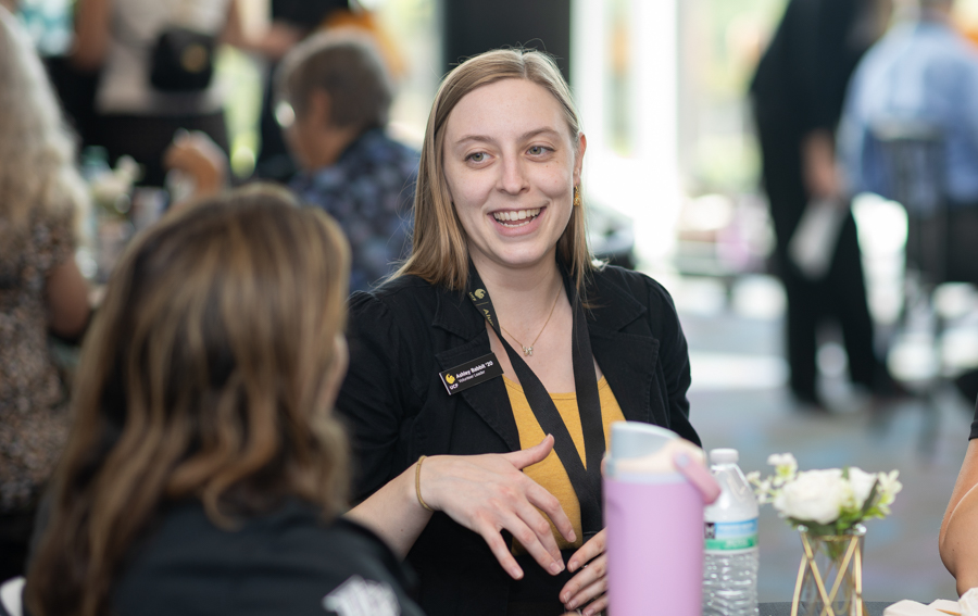 Smiling woman with a lanyard badge speaks with another attendee at a conference table during a networking event.