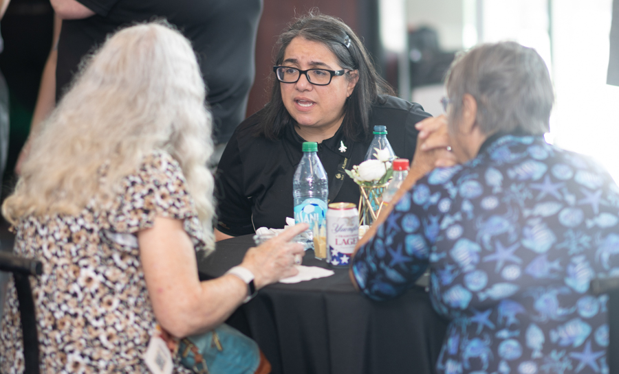 Woman with glasses explains something to two older women seated at a round table with water bottles and papers in a bright room.