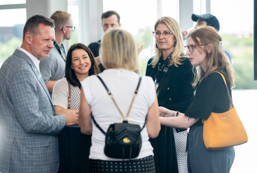 Group of professionals chatting in a bright hallway at a networking event; diverse men and women wearing business casual attire with lanyards.