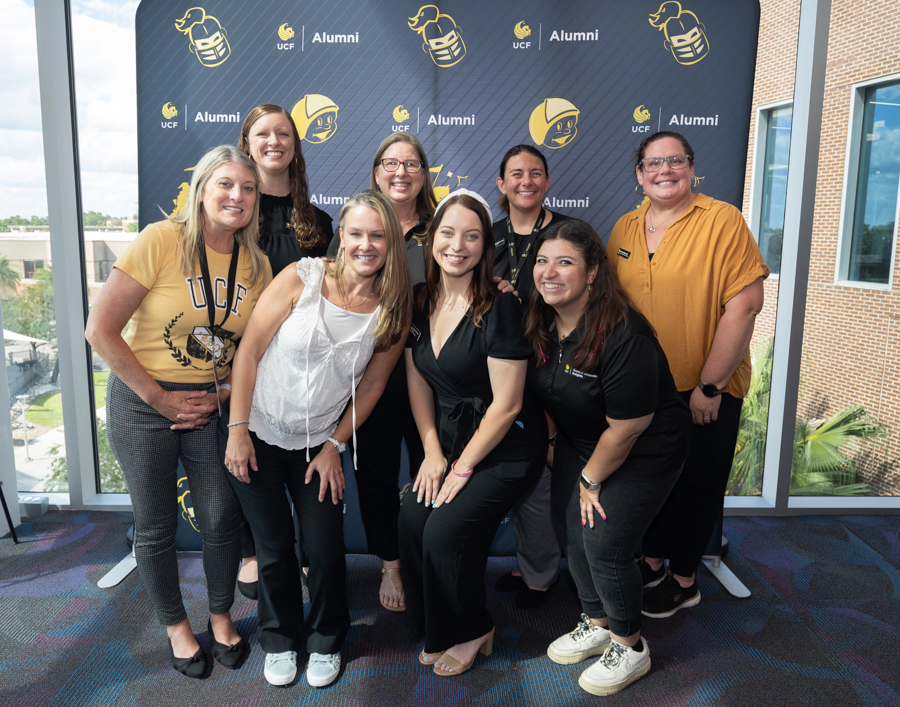 Eight women posing for a group photo at a UCF Alumni event in front of a branded backdrop.
