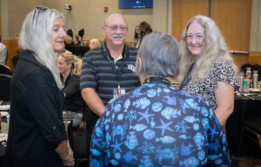 Group of four adults standing in a conference room, conversing near a round table with black tablecloths and place settings, wearing name badges and lanyards.