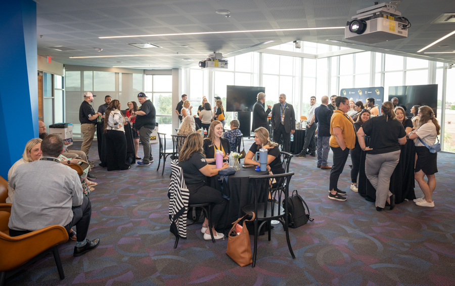 Professionals mingle at a corporate networking event in a bright, modern venue with round tables and attendees chatting nearby.
