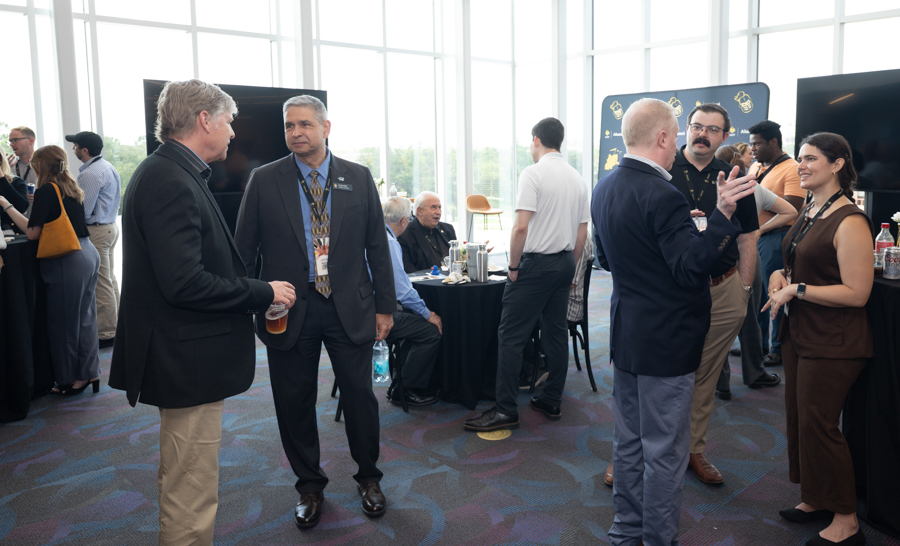 Professionals conversing at a networking event in a bright atrium, some holding drinks and wearing name badges around tables with attendees in the background.