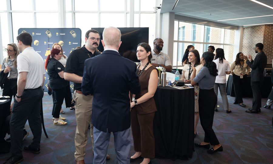 Group of professionals networking at a conference reception, chatting near a high-top table with a backdrop banner and a large screen in a bright venue.
