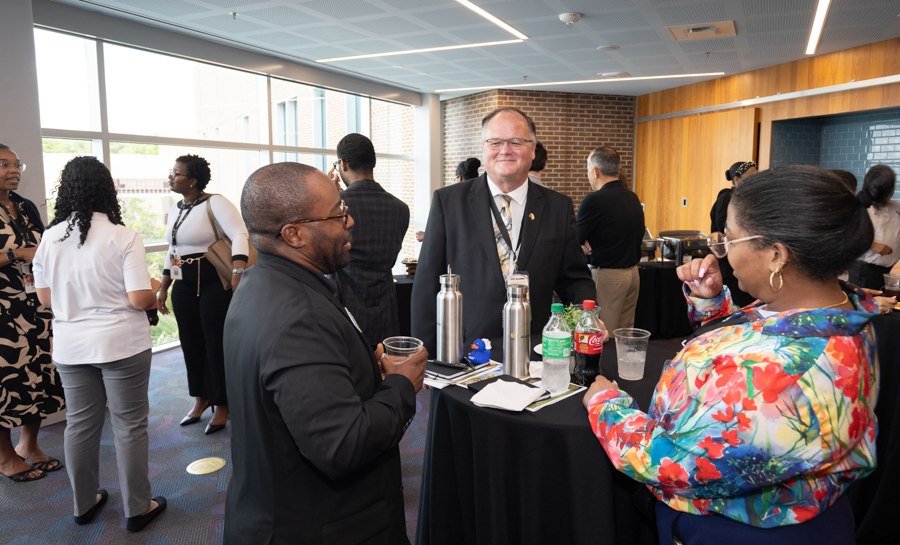 Professionals chat around a high round table with coffee pots and soda bottles at a daytime networking reception in a modern venue.