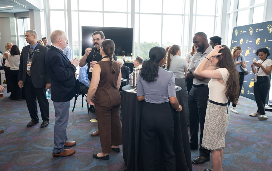 Group of professionals socializing at a networking event in a modern, bright venue with tall round tables and large windows.