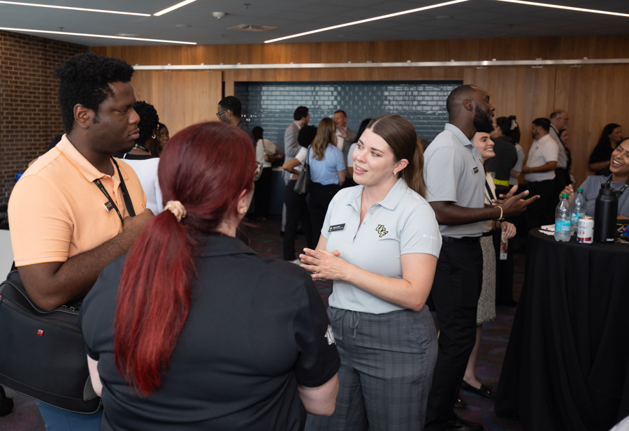 Professionals networking in a modern venue; a woman in a light blue polo chats with a man in an orange shirt as others mingle nearby.