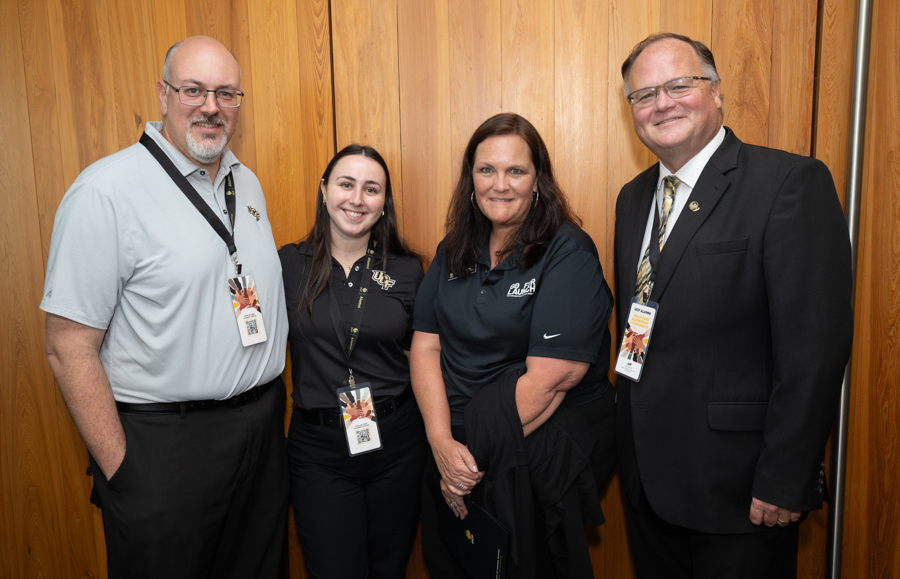 Four adults posing for a photo in a wood-paneled room, wearing lanyards with badges and smiling.