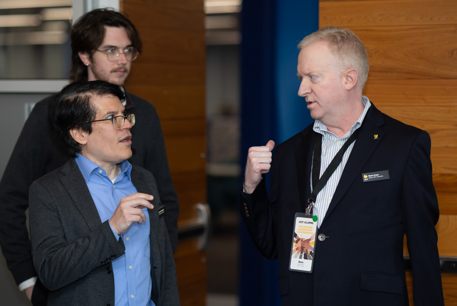 Three men in business attire converse in a modern hallway; the man in a blue shirt and glasses gestures while the other listens, wearing a dark blazer and lanyard.