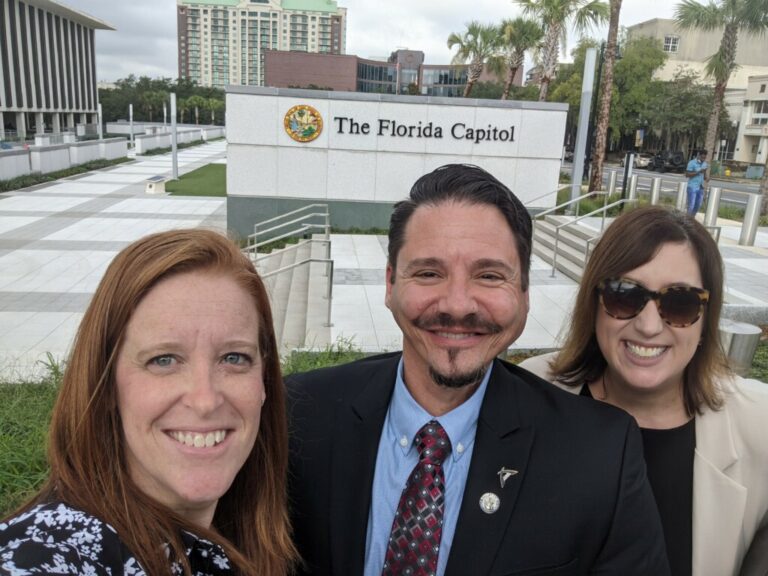 Three people smiling for a selfie outside The Florida Capitol sign, with palm trees and buildings in the background.