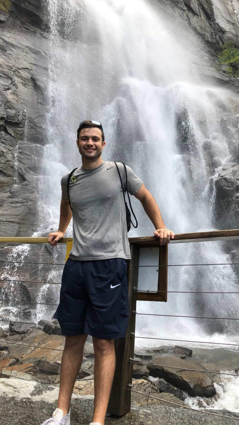 Man standing at a railing in front of a large waterfall, smiling, wearing a gray Nike t‑shirt, navy shorts, backpack, and sunglasses on his head.