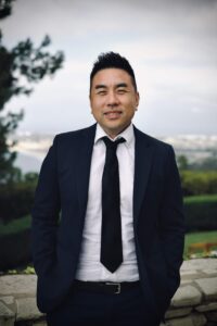 Smiling man in a navy suit and black tie standing outdoors with blurred trees and cityscape in the background.