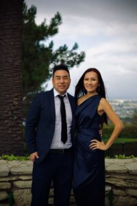 A couple in formal attire posing outdoors, man in a navy suit and tie, woman in a dark blue evening gown, with a palm tree behind them.