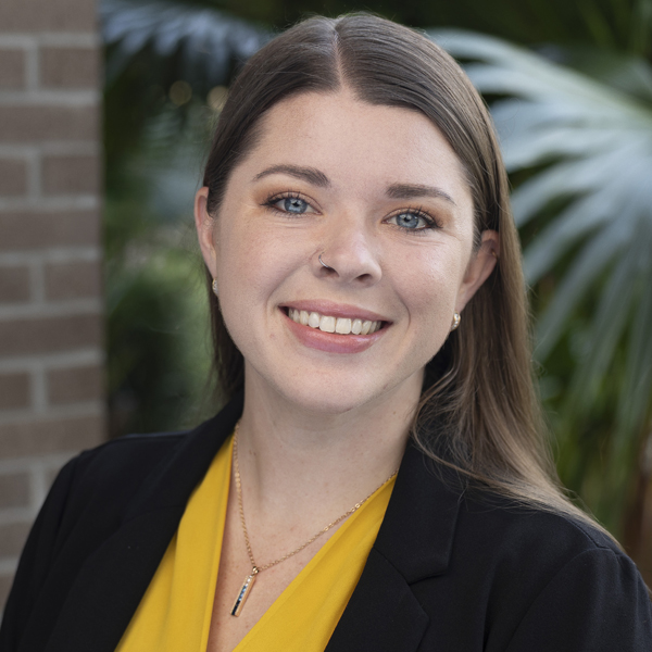 Portrait of a smiling woman with light brown hair in a black blazer and yellow blouse, outdoors.