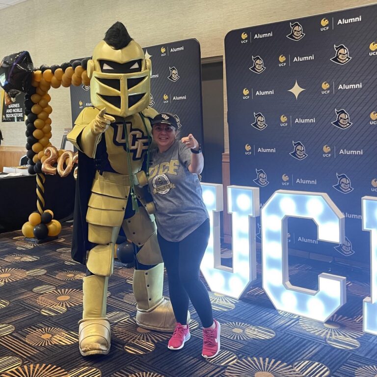 Person posing with a gold knight mascot at a UCF Alumni event, yellow/black balloon arch in the background.
