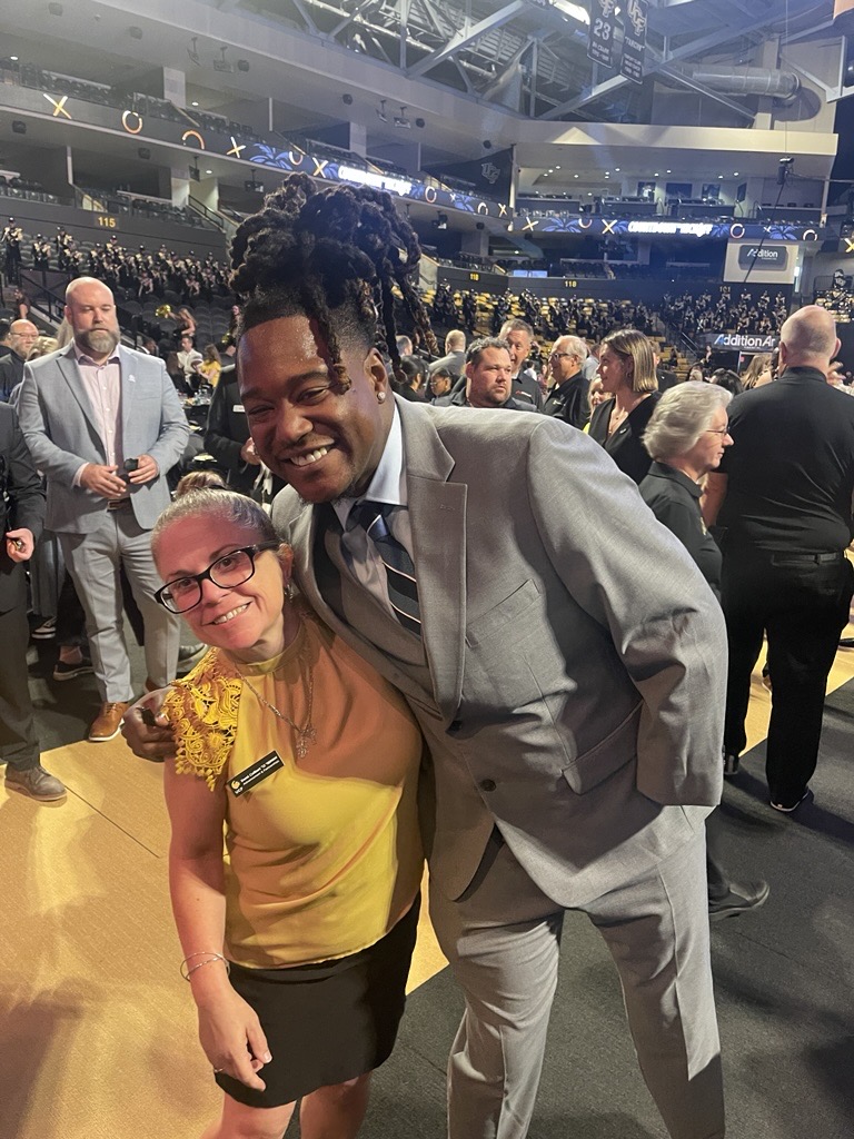 Two people smile at the camera: a tall man in a light gray suit leaning over a woman in a yellow top and black skirt, in a crowded indoor venue with many attendees in the background.