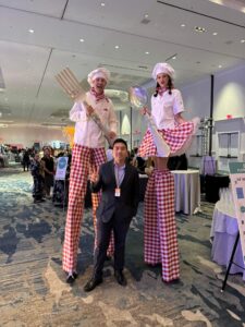 Two tall stilt-walkers dressed as chefs in white uniforms and red-checkered pants pose with a man at an indoor event hall, holding oversized utensils (a spatula and a ladle).
